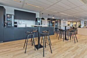 Moderne bedrijfskantine met zwarte keuken, barkrukken en hoge tafels op een houten vloer aan de Prof. W.H. Keesomlaan.